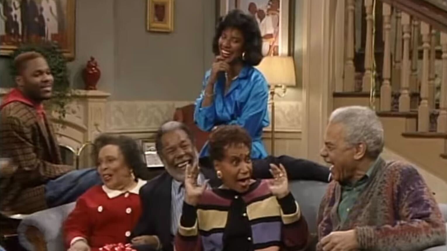 The cast of The Cosby Show sitting together in the Huxtable family living room.