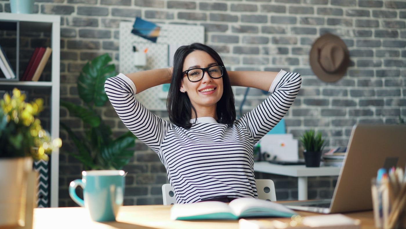 Woman in glasses and a striped top leans back at her desk with hands behind her head, smiling relaxed with a laptop, mug, and notebook in front of her.