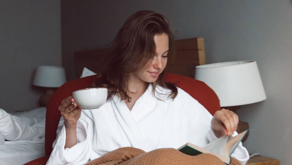 A lady in a white bathrobe sits on a cozy red chair, holding a white cup as she reads a book. A brown blanket is draped over her lap, and soft lighting, bedside lamps, and an unmade bed create a calm atmosphere that suggests an evening indoors.