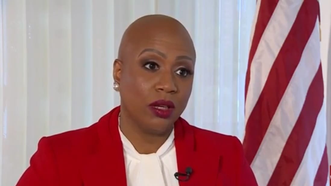 Close-up portrait of Rep. Ayanna Pressley during a television interview, wearing a red blazer and white blouse, with a U.S. flag visible in the background.