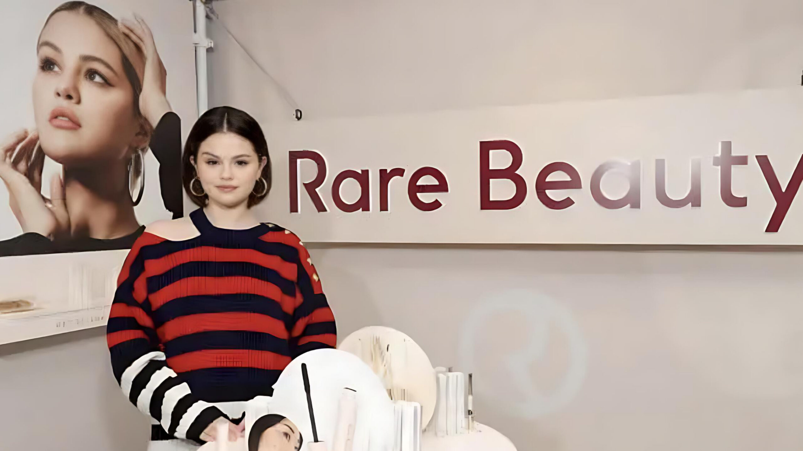 Selena Gomez standing in front of a Rare Beauty sign, wearing a red and navy striped off-shoulder sweater, with makeup products displayed on a table in front of her.