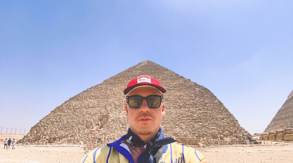 Macklemore wearing a red cap, sunglasses, and a neck bandana posing for a selfie in front of the Great Pyramid of Giza