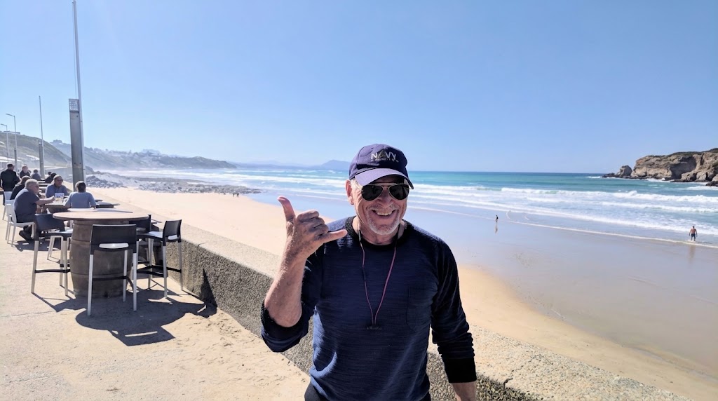 Musician Jimmy Buffett smiling and making a shaka hand sign on a beach boardwalk.
