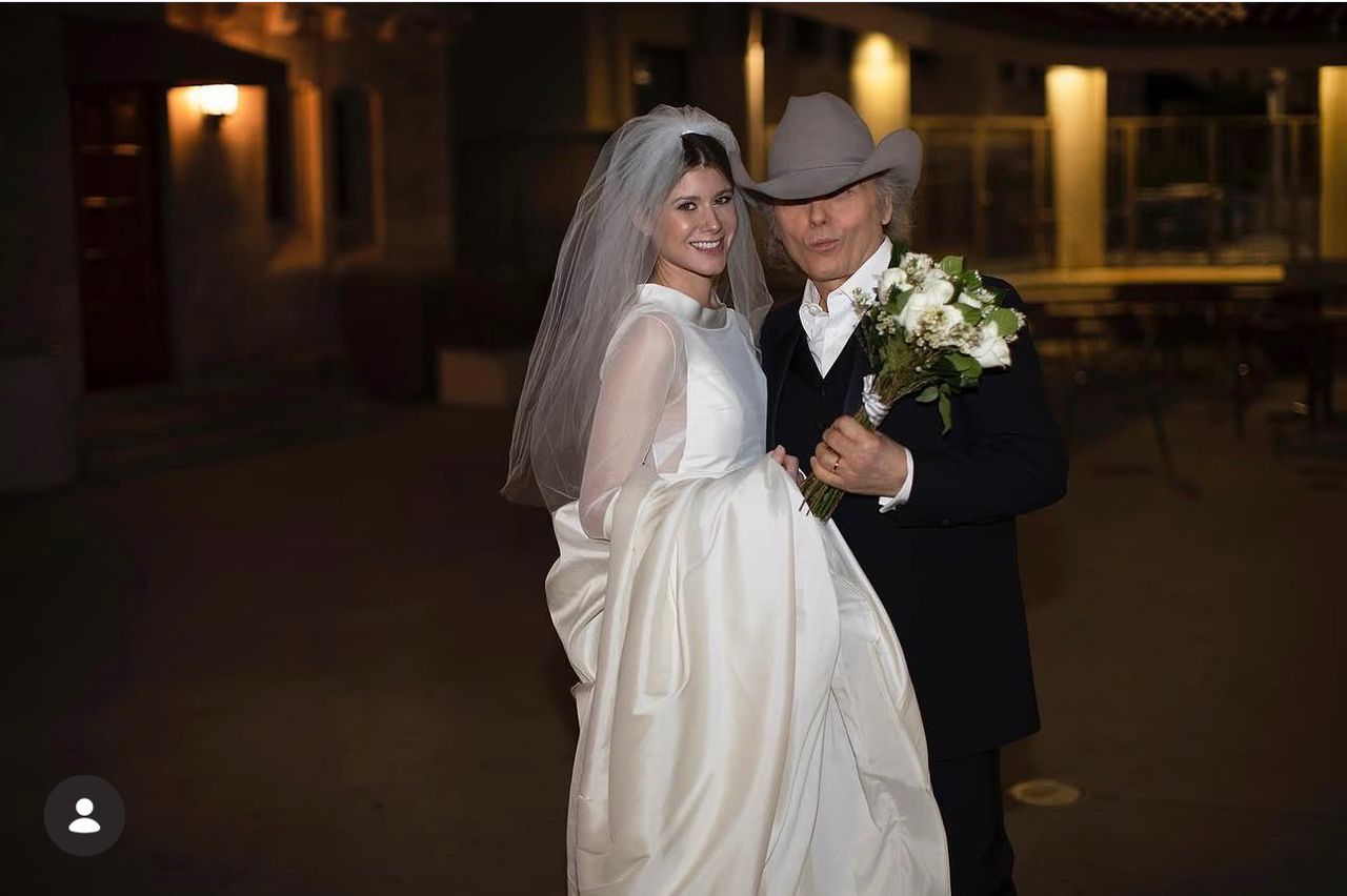 Dwight Yoakam and his wife pose together at their wedding, with Yoakam in a cowboy hat and his wife in a white wedding dress and veil.