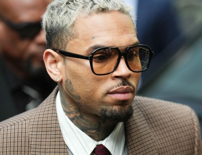 Chris Brown looking forward while wearing tinted aviator glasses, a patterned brown suit jacket, and displaying visible neck and face tattoos.