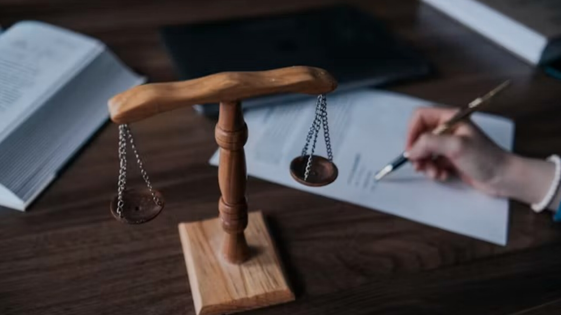 Close-up of a wooden desk featuring a small decorative balance scale in the foreground. In the soft-focus background, a person holds a metallic fountain pen, signing a paper document beside open legal books.