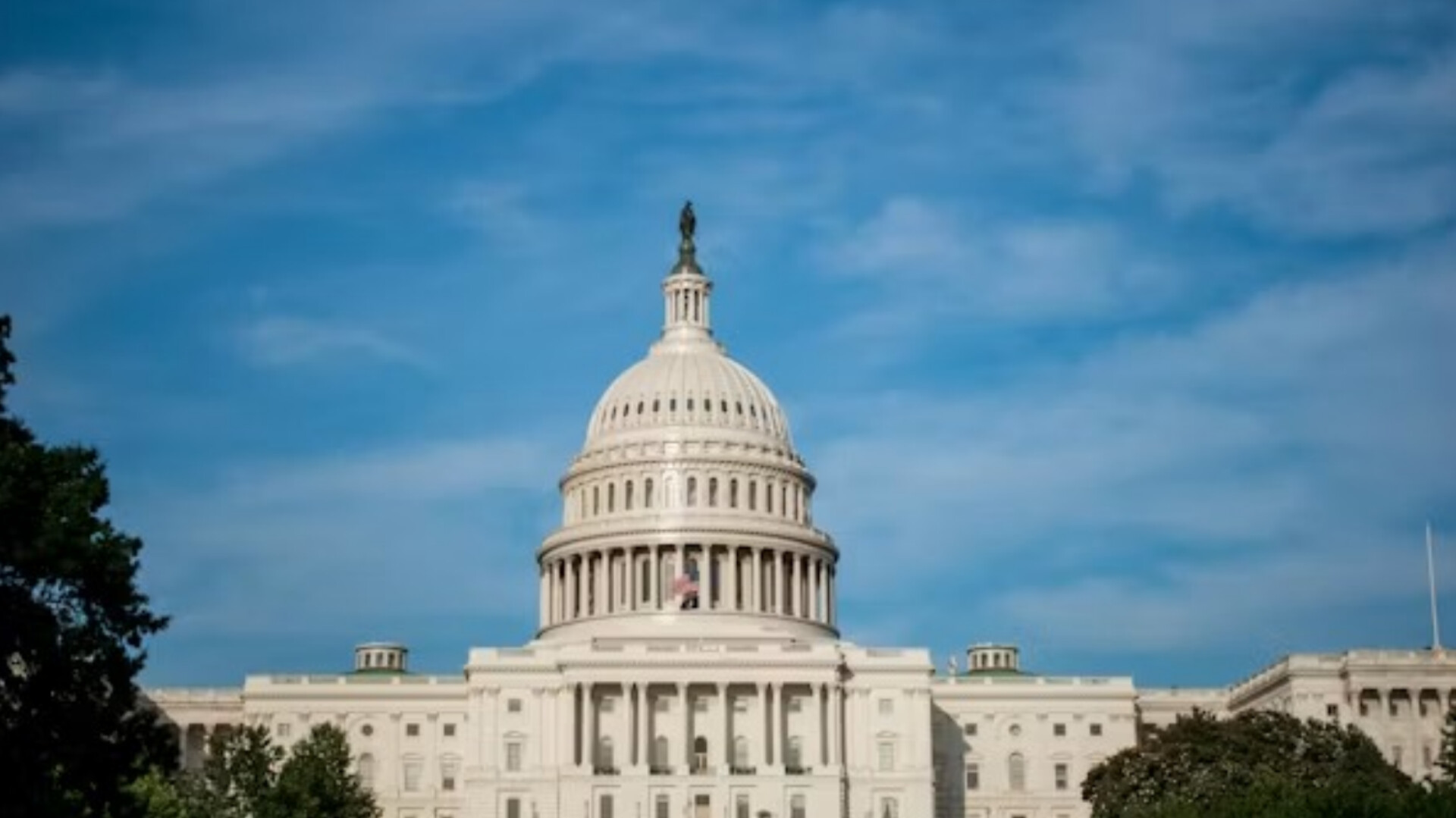 Front-facing shot of the U.S. Capitol dome and upper building in Washington, D.C., constructed in white marble, set against a partly cloudy blue sky with green tree tops visible at the bottom corners.