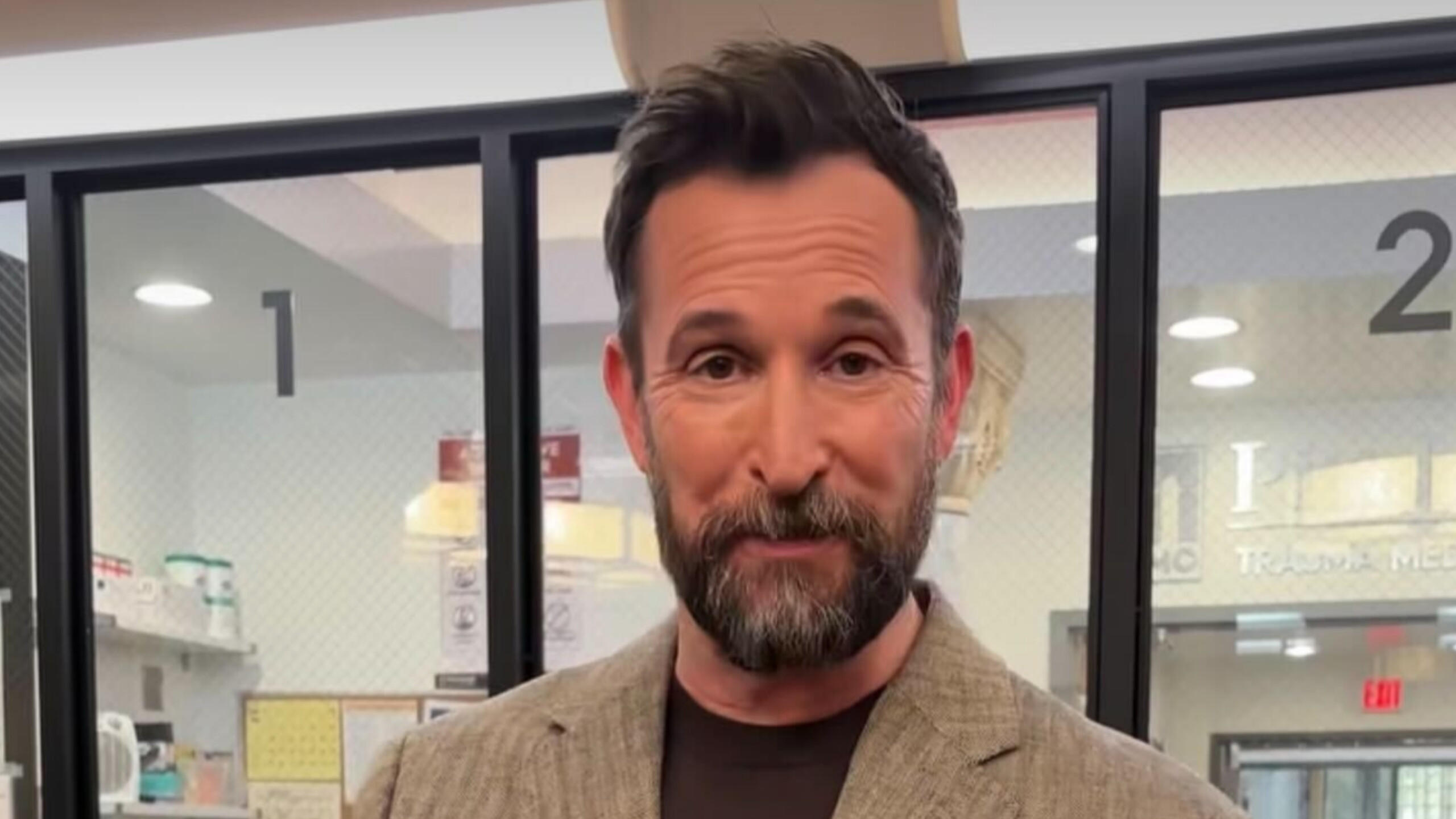 Actor Noah Wyle looking into the camera with a beard and beige jacket.
