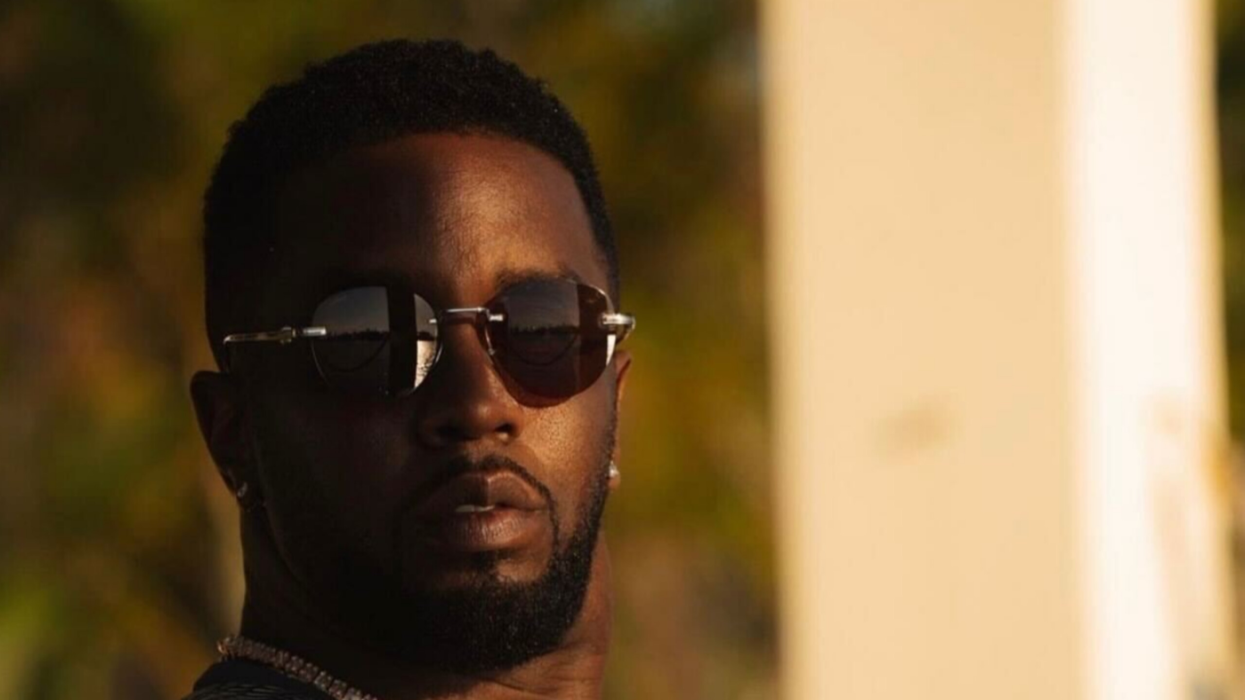 Sean "Diddy" Combs wearing dark rimless sunglasses and a silver chain necklace in warm sunlight.
