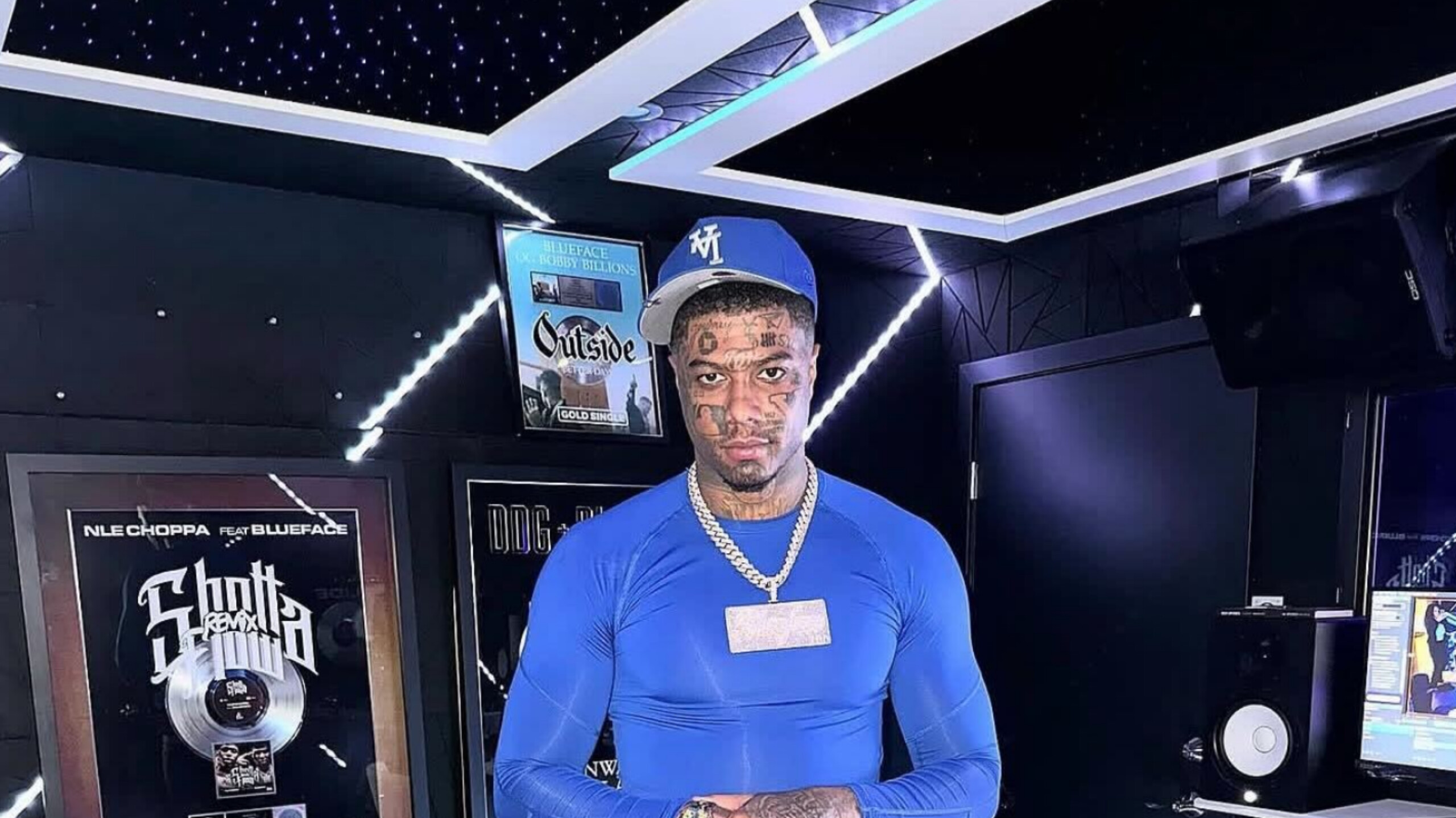 Rapper Blueface standing in a music recording studio wearing a royal blue long-sleeve shirt and matching cap.
