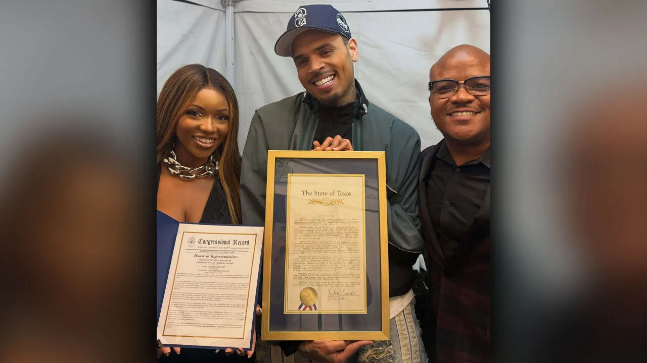Three people stand together smiling for a photo. On the left, Rep. Jasmine Crockett holds a framed Congressional Record document. In the center, singer Chris Brown, wearing a navy baseball cap and jacket, holds a large gold-framed resolution from "The State of Texas." On the right, a smiling man in a black button-up shirt poses alongside them.