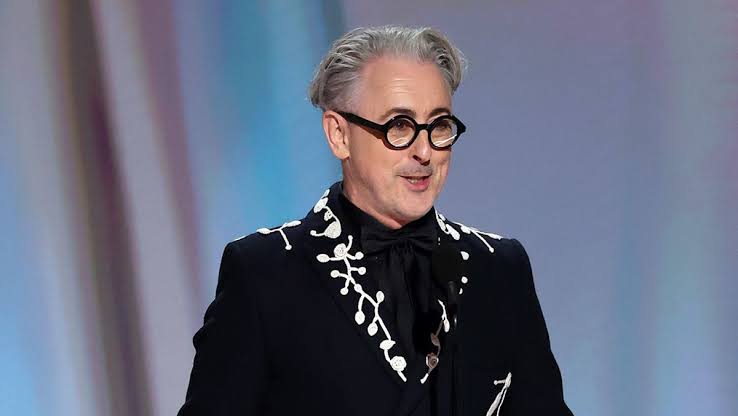 Medium close-up of Alan Cumming on stage with slicked-back gray hair and round black-rimmed glasses. He is wearing a black suit jacket styled with intricate white vine-like embroidery along the lapels, paired with a black shirt and bow tie, set against a muted pastel-toned backdrop.