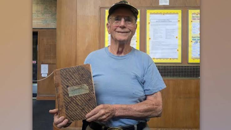 Medium portrait of an elderly man wearing a blue t-shirt, glasses, and a dark cap, smiling at the camera. He is holding a heavily worn, vintage book with a marbled brown cover in front of a wooden wall with yellow bulletin boards.