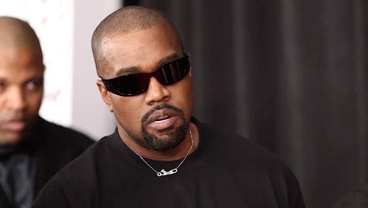 Kanye West wearing dark sunglasses and a black t-shirt while speaking.