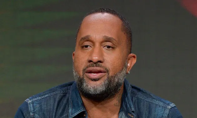 Close-up portrait of writer and producer Kenya Barris looking slightly off-camera with a neutral expression while speaking. He has a trimmed graying beard, short dark hair, a small earring, and is wearing a dark blue denim jacket against a dark background.