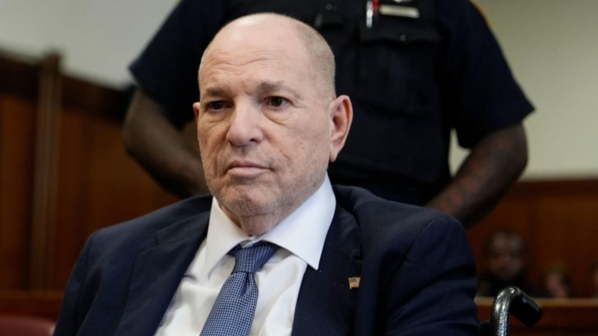 Harvey Weinstein sitting in a suit inside a courtroom with a law enforcement officer standing behind him.