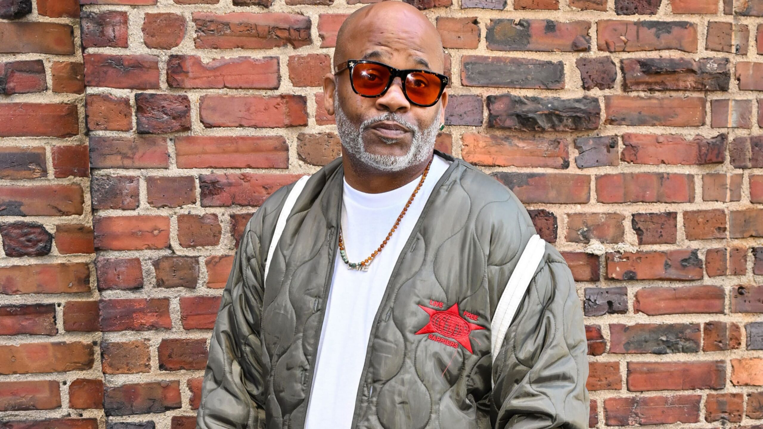 Record executive Dame Dash wearing tinted sunglasses, a bead necklace, and an olive green quilted jacket posing in front of a red brick wall.