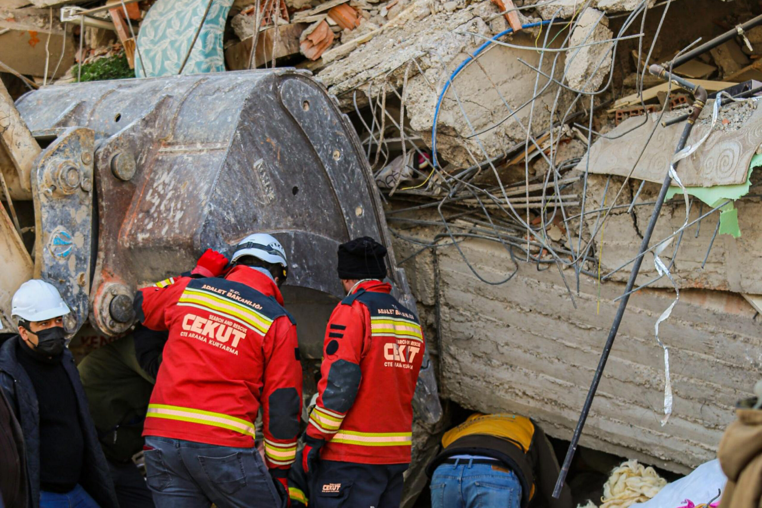 Rescue workers in red CEKUT search-and-rescue jackets stand beside a large excavator bucket amid the rubble of a collapsed building, with concrete debris, twisted rebar, and other responders nearby.