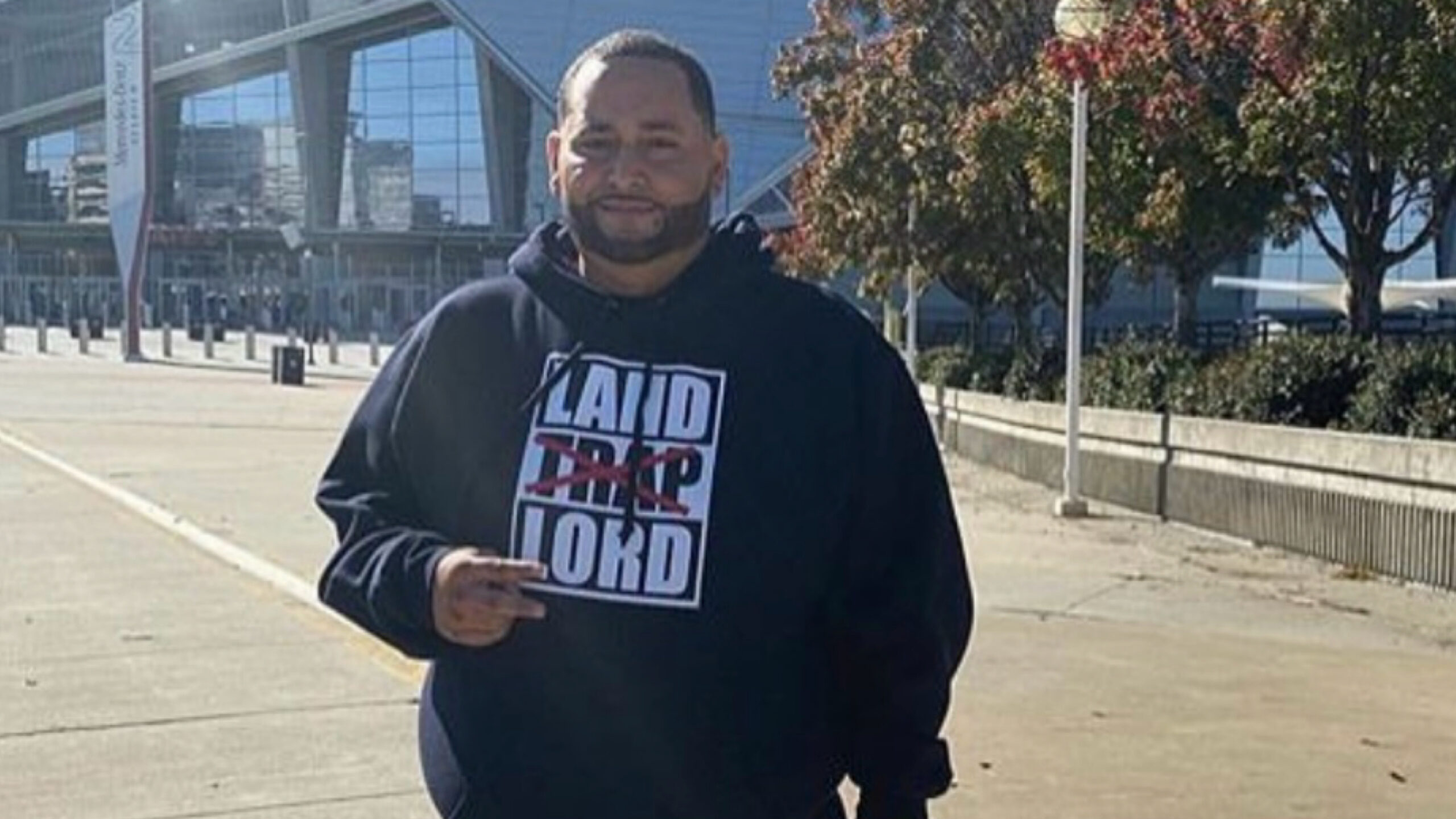 Cesar Pina wearing a black "LAND LORD" graphic hoodie standing on a paved walkway outside a stadium concourse.