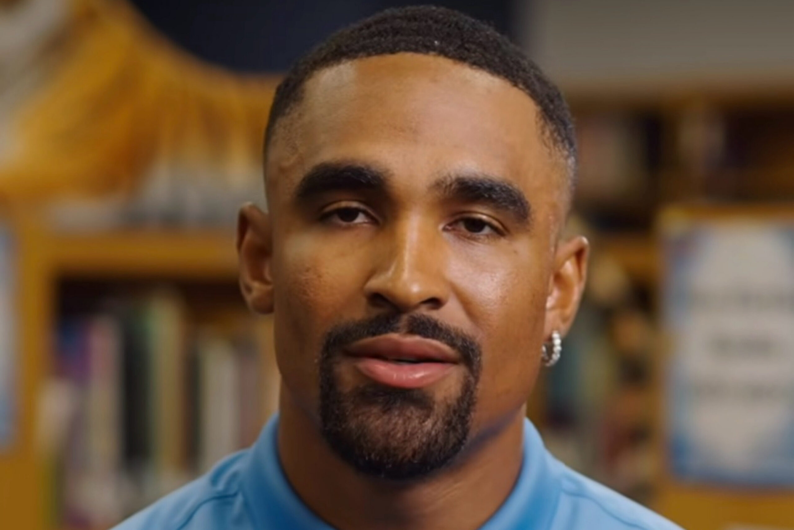 Actor Jalen Hurts / educator character closeup looking toward the camera wearing a light blue polo shirt and a small hoop earring indoors.