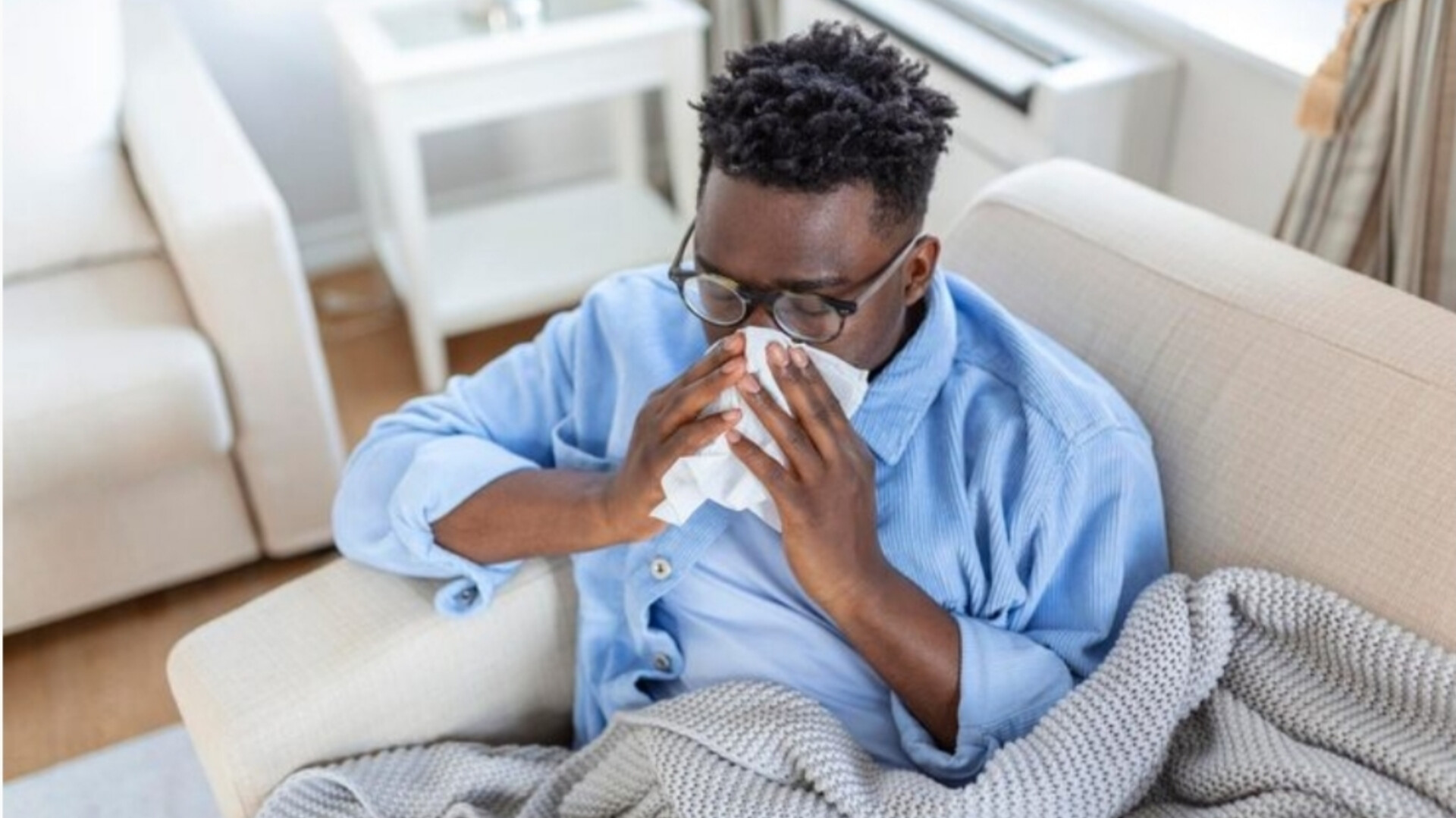 A man blowing his nose with a tissue while wrapped in a blanket on a couch.