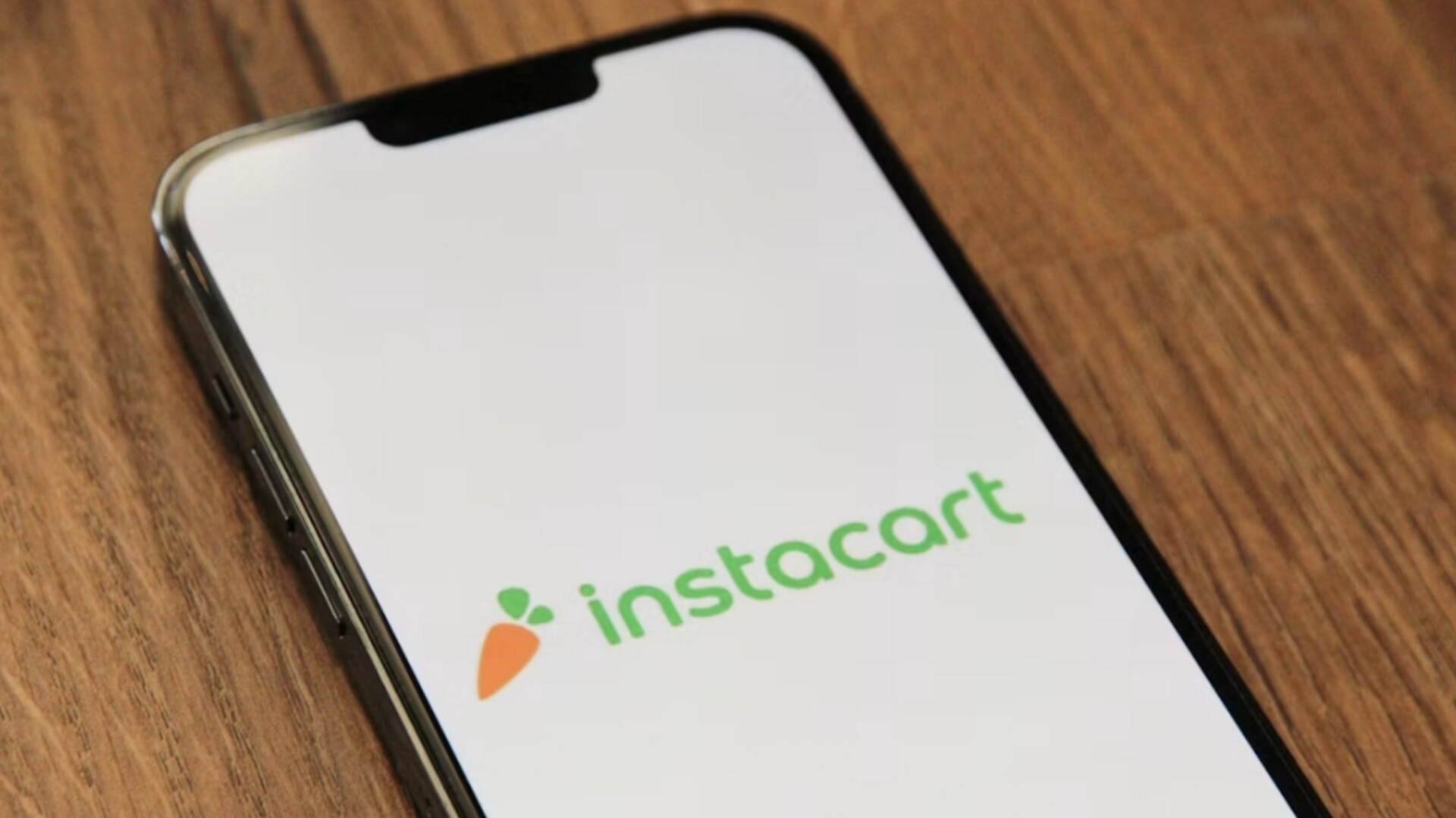 A smartphone lying on a wooden surface displaying the Instacart logo on its screen.