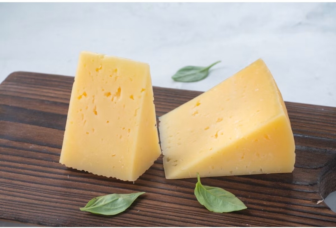 Two triangular blocks of yellow cheese resting on a dark wooden cutting board with fresh green leaves.