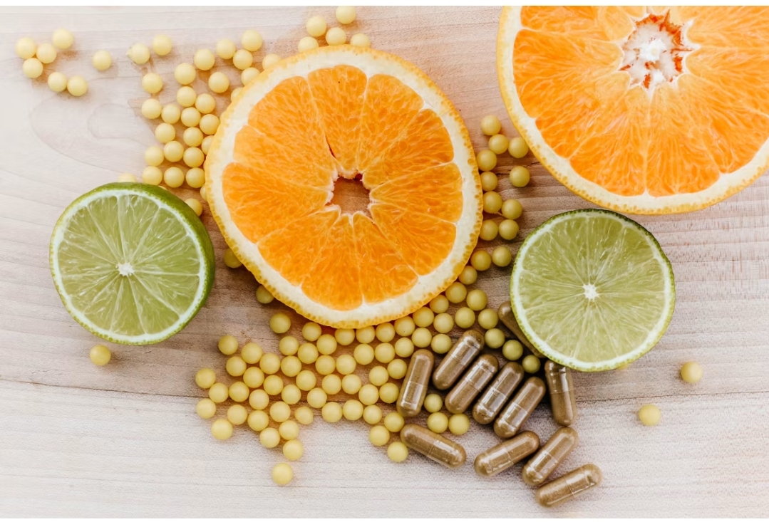 Citrus fruit slices, yellow Vitamin C pellets, and brown supplement capsules arranged on a light wooden background.
